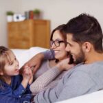Cute little girl in blue denim dress with folded hands and laughing in parents lap while they lay back in comfortable sofa indoors